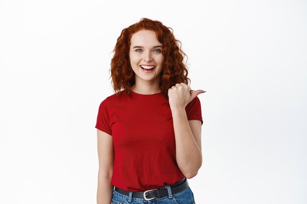 Portrait d'une belle femme rousse heureuse montrant, pointant vers la droite et souriante joyeuse, recommandant la compagnie, mur blanc