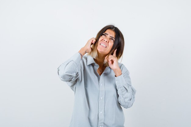 Portrait d'une belle femme se branchant les oreilles avec les doigts, levant les yeux en chemise et regardant la vue de face effrayée