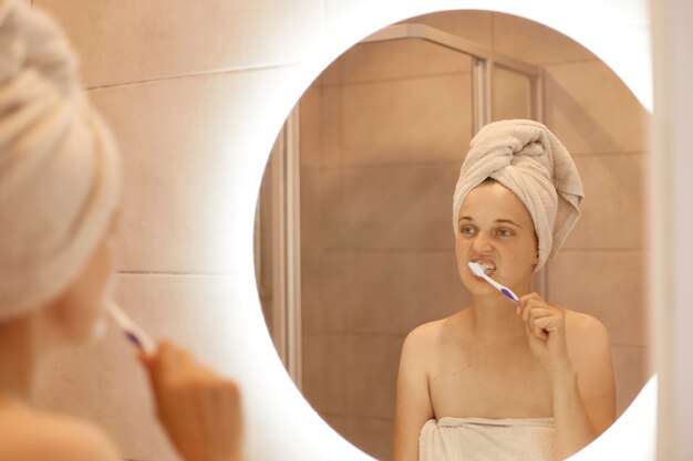 Portrait d'une belle femme séduisante avec une serviette sur la tête, debout devant le miroir de la salle de bain et se brossant les dents, procédures d'hygiène le matin.