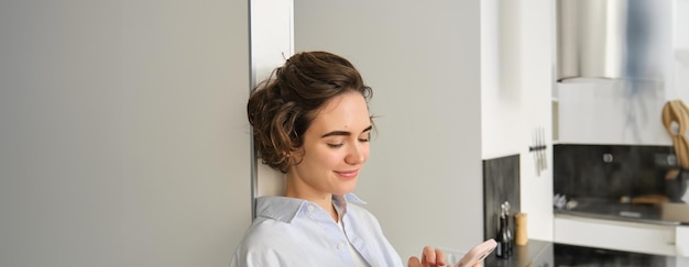 Photo gratuite portrait d'une belle femme souriante regardant un smartphone debout à la maison près de la cuisine en train de vérifier