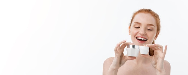 Portrait d'une belle femme souriante tout en prenant de la crème faciale isolée sur fond blanc avec