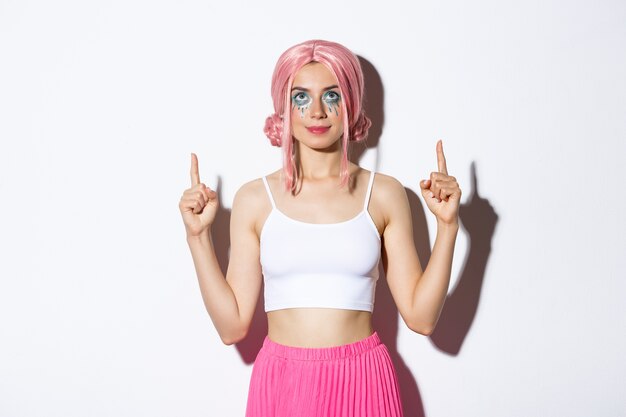 Portrait de belle fêtarde avec perruque rose et maquillage glamour, pointant du doigt et levant les yeux, montrant votre bannière de logo, debout.