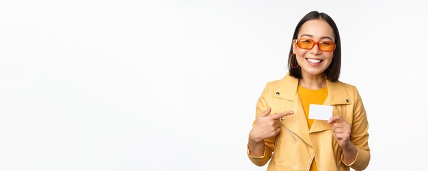 Portrait de belle fille asiatique moderne à lunettes de soleil souriant heureux montrant la carte de crédit debout sur l'espace de copie de fond blanc