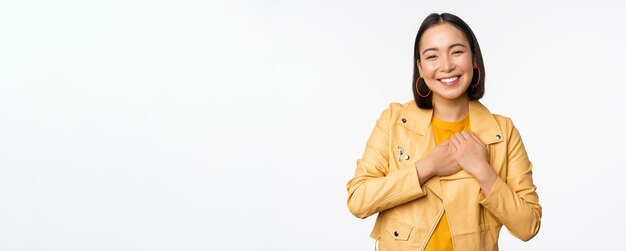Portrait d'une belle fille asiatique souriante regardant avec appréciation merci geste tenant la main sur le cœur flatté debout sur fond blanc