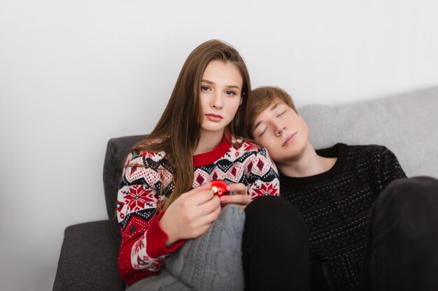 Portrait d'une belle fille assise sur un canapé et regardant rêveusement à huis clos avec un jouet de Noël à la main pendant que le garçon s'appuie sur elle et dort