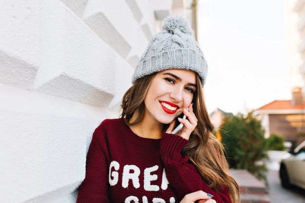 Portrait belle fille aux cheveux longs en pull marsala parlant au téléphone dans la rue. Elle porte un bonnet tricoté et sourit.