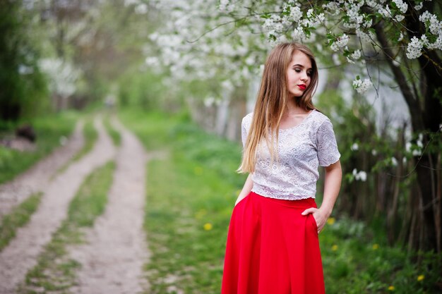 Portrait d'une belle fille aux lèvres rouges au jardin de fleurs de printemps portant une robe rouge et un chemisier blanc