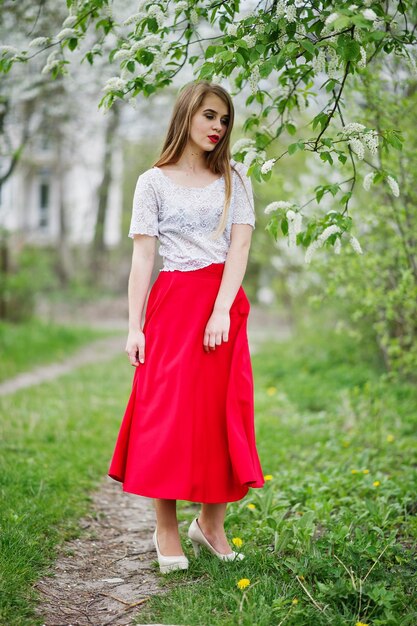 Portrait d'une belle fille aux lèvres rouges au jardin de fleurs de printemps portant une robe rouge et un chemisier blanc