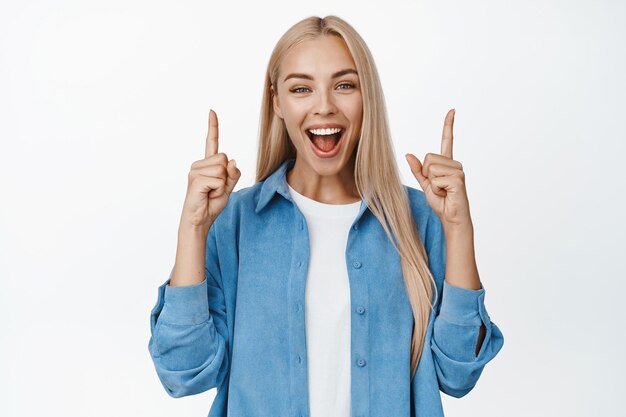 Portrait d'une belle fille blonde faisant une annonce pointant les doigts vers le haut et souriant à la caméra montrant le fond blanc de la publicité