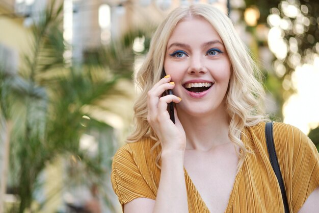 Portrait d'une belle fille blonde joyeuse parlant joyeusement sur un téléphone portable en plein air
