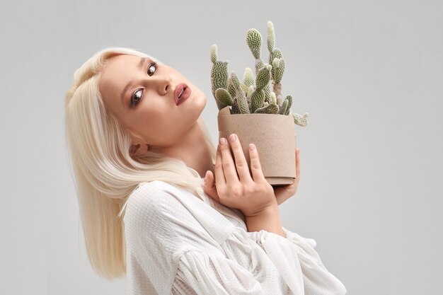 Portrait d'une belle fille blonde avec maquillage et coiffure portant un chemisier blanc, regardant la caméra et tenant un pot avec un cactus vert. Jolie jeune femme achetant l'usine pour la maison