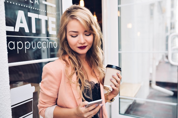 Portrait belle fille blonde sur terrasse avec une tasse de café. Elle porte une veste corail, des lèvres vineuses, souriant au téléphone à la main.