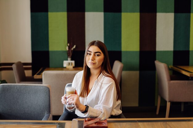 Portrait de belle fille, boire du thé chaud ou du café dans un café avec son téléphone portable.