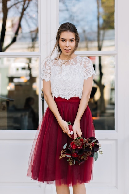 Portrait belle fille avec une coiffure légère en jupe de tulle marsala sur rue. Elle tient le bouquet de fleurs et à la recherche