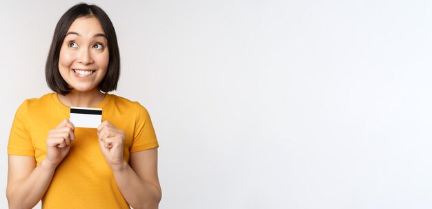 Portrait d'une belle fille coréenne tenant une carte de crédit recommandant un service bancaire debout en t-shirt jaune sur fond blanc