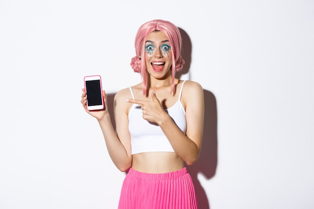 Portrait de belle fille excitée en costume d'halloween, perruque rose et maquillage lumineux, pointant le doigt sur un téléphone mobile avec un visage étonné, debout.