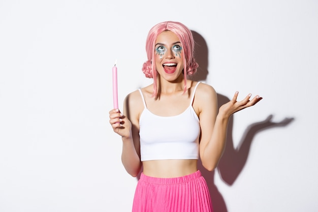 Photo gratuite portrait de belle fille excitée déguisée en fée à l'halloween, portant une perruque rose et un maquillage lumineux, tenant une bougie et parlant à quelqu'un, debout.