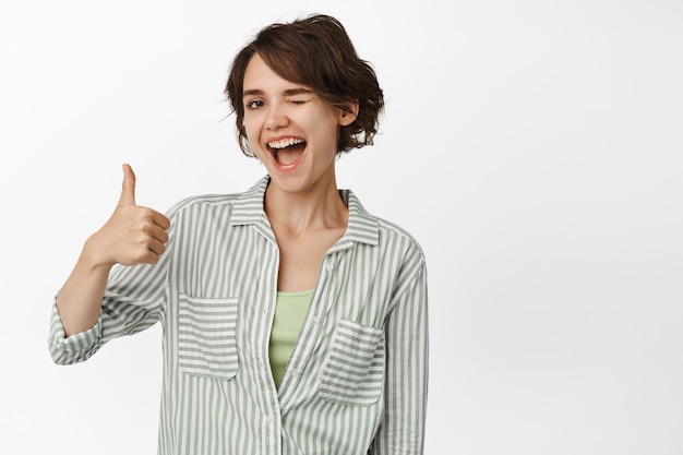 Portrait d'une belle fille faisant un clin d'œil et souriant, montrant les pouces vers le haut, des commentaires positifs, assure que tout va bien, très bien, loue un bon travail, debout sur blanc