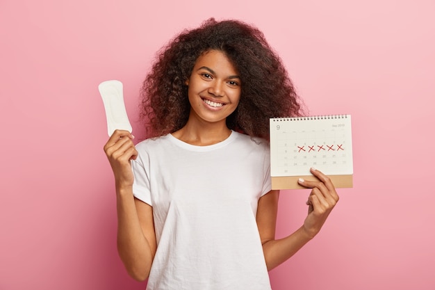 Portrait de belle fille féminine a une coiffure afro, tient un calendrier mensuel, une serviette hygiénique propre