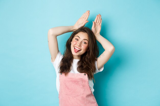 Portrait d'une belle fille glamour célébrant Pâques, montrant des oreilles de lapin avec les mains au-dessus de la tête, souriante joyeuse, debout sur fond bleu.