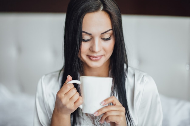 Portrait d'une belle fille heureuse brune assise dans son lit le matin et buvant du thé dans la tasse mignonne