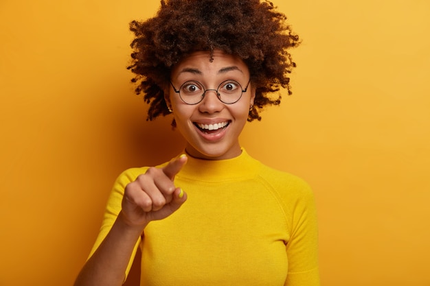 Portrait de belle fille heureuse de sexe féminin pointe le doigt avant, choisit quelque chose devant, sourit joyeusement, porte des vêtements décontractés, pose contre le mur jaune. Je te sélectionne, rejoins moi