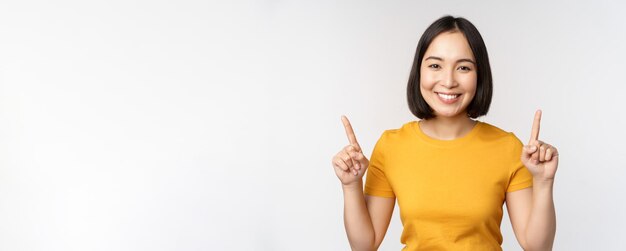 Portrait d'une belle fille japonaise souriante pointant les doigts vers le haut montrant une publicité debout en t-shirt jaune sur fond blanc