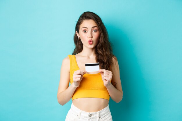 Portrait d'une belle fille surprise qui a l'air amusée et qui plisse les lèvres, montrant une carte de crédit en plastique, debout sur fond bleu. Espace de copie