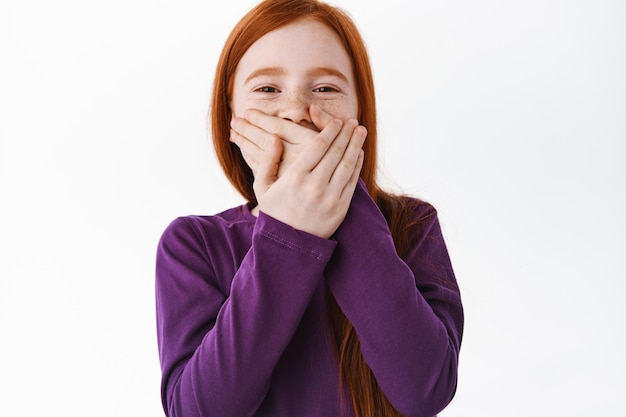 Photo gratuite portrait d'une belle fille à tête rouge avec des taches de rousseur riant, couvrant sa bouche avec la main et riant sur une chose drôle, plaisantant, debout sur fond blanc