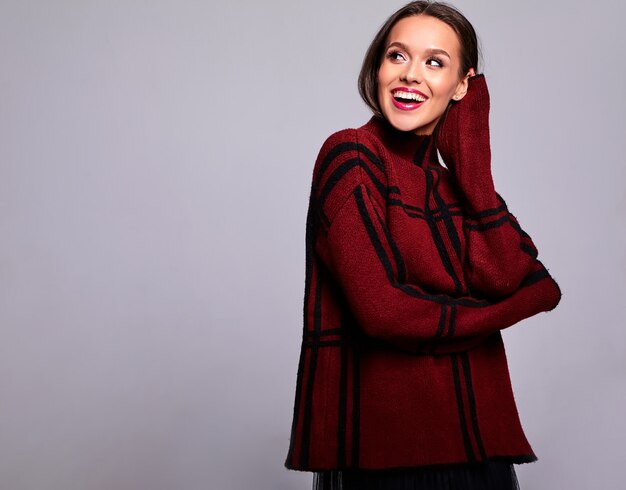 Portrait de la belle heureuse femme brune mignonne modèle dans des vêtements de chandail rouge chaud décontracté isolé sur gris avec du maquillage de soirée et des lèvres colorées