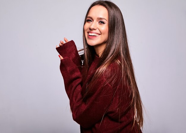 Portrait de la belle heureuse femme brune mignonne modèle dans des vêtements de chandail rouge chaud décontracté isolé sur gris avec du maquillage de soirée et des lèvres colorées