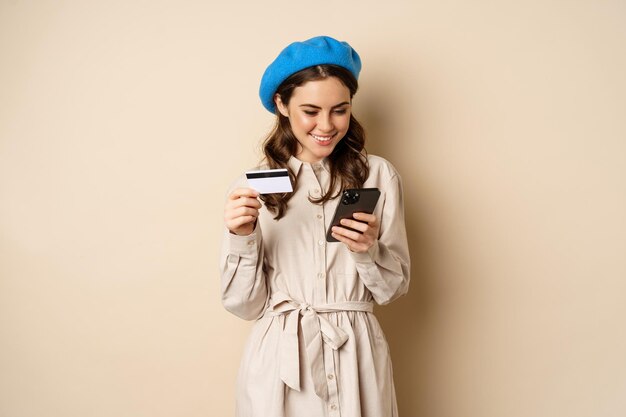 Portrait d'une belle jeune femme de 25 ans utilisant une application d'achat de téléphone portable, une carte de crédit, une livraison de commande, un achat, debout sur un fond beige