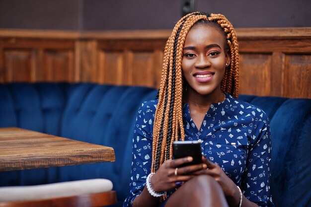 Portrait de la belle jeune femme d'affaires africaine avec des dreadlocks porter sur un chemisier bleu et une jupe assis dans un café avec un téléphone portable