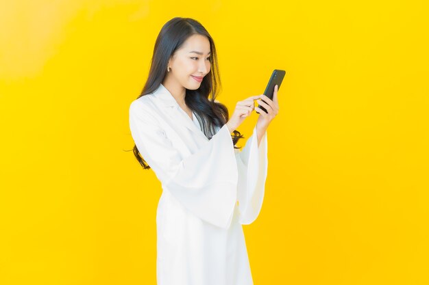 Portrait d'une belle jeune femme asiatique souriante avec un téléphone portable intelligent sur un mur jaune