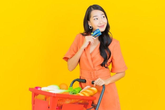 Portrait belle jeune femme asiatique sourire avec panier d'épicerie de supermarché sur jaune