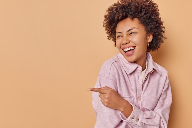 Photo gratuit portrait d'une belle jeune femme aux cheveux bouclés ravie qui rit joyeusement et pointe vers l'espace de copie vierge montre quelque chose de drôle porte une veste isolée sur fond beige. ha ha regarde ça