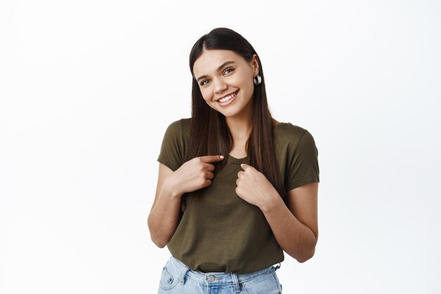 Portrait de la belle jeune femme en bonne santé en t-shirt pointant vers elle-même et souriant timide autopromotion demandant de la choisir debout sur fond blanc