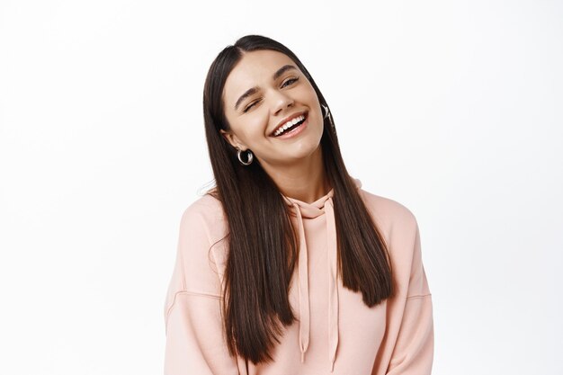 Portrait d'une belle jeune femme candide clin d'œil à l'avant, souriante et riant, debout heureuse en sweat à capuche contre un mur blanc, restant positive