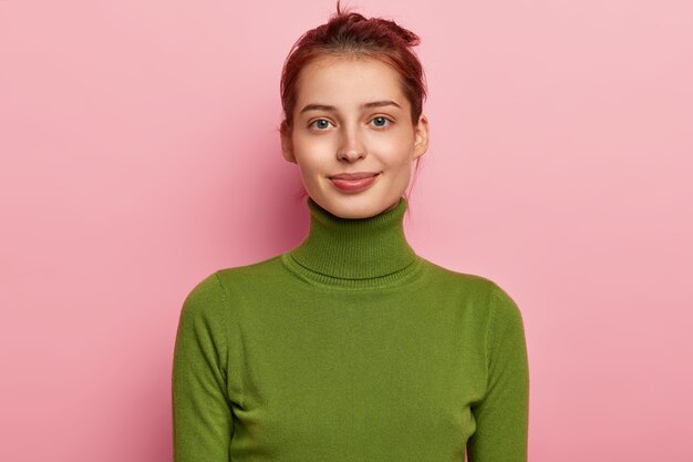 Portrait de la belle jeune femme a les cheveux peignés foncés, apparence attrayante, porte un col roulé vert décontracté, regarde volontiers dans la caméra, pose sur fond rose