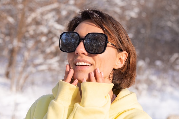 Photo gratuite portrait d'une belle jeune femme dans une forêt d'hiver paysage enneigé sur une journée ensoleillée, vêtue d'un gros pull jaune, avec des lunettes de soleil, profitant du soleil et de la neige