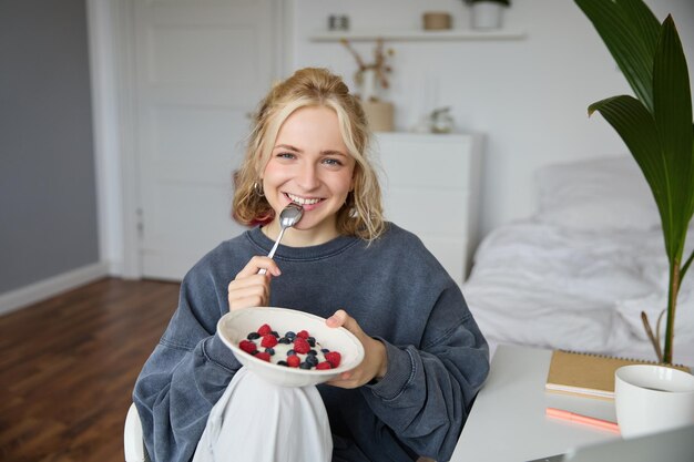 Portrait d'une belle jeune femme dans une pièce mangeant le petit déjeuner tenant un bol avec un dessert et une cuillère