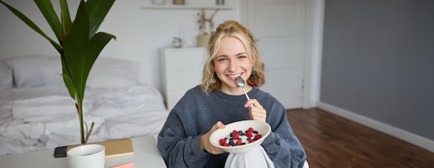 Portrait d'une belle jeune femme dans une pièce mangeant le petit déjeuner tenant un bol avec un dessert et une cuillère