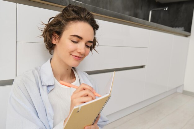 Portrait d'une belle jeune femme écrivant dans un journal en ajoutant des notes dans le planificateur assis sur le sol et th