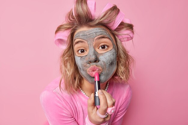 Portrait d'une belle jeune femme européenne applique un masque nourrissant de beauté rouge à lèvres et des bigoudis se préparent pour la date veut être belle utilise des produits cosmétiques isolés sur fond rose