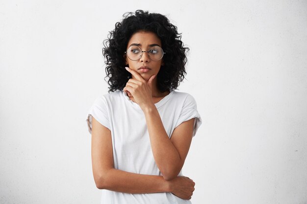 Portrait de la belle jeune femme habillée avec désinvolture à lunettes rondes ayant une expression douteuse