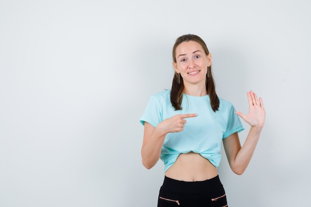 Portrait d'une belle jeune femme pointant vers la droite, agitant la main pour saluer en t-shirt, pantalon et à la vue de face béate