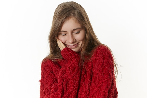 Portrait de belle jeune femme de race blanche positive avec un sourire amical heureux, gardant les yeux fermés, rêvant, touchant la joue, portant un pull marron chaud à la mode sur une froide journée d'hiver