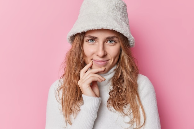 Portrait d'une belle jeune femme regarde directement la caméra avec une expression satisfaite sourit agréablement porte un panama blanc et un col roulé isolé sur fond rose écoute des nouvelles positives