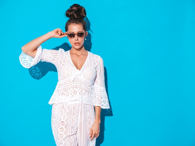 Portrait de la belle jeune femme sexy avec une coiffure de goule. Fille à la mode dans des vêtements de costume hipster blanc d'été décontracté dans des lunettes de soleil. Modèle chaud isolé sur bleu