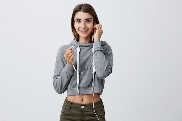 Portrait De La Belle Jeune Fille étudiante Caucasienne Attrayante Aux Cheveux Longs Noirs En Tenue élégante Décontractée Souriant Brillamment, écoutant La Musique Préférée Sur Le Chemin De L'université. Concept De Style De Vie Moderne.